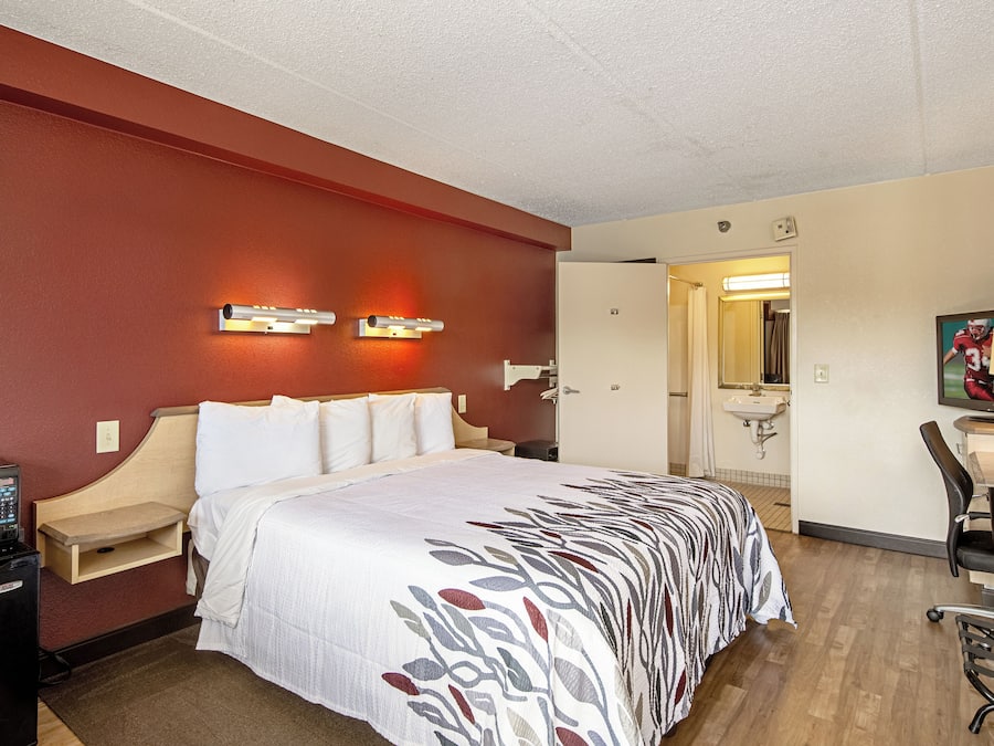Quarto de hotel com cama, mesa de cabeceira, luminária, escrivaninha, cadeira e banheiro visível por uma porta aberta.