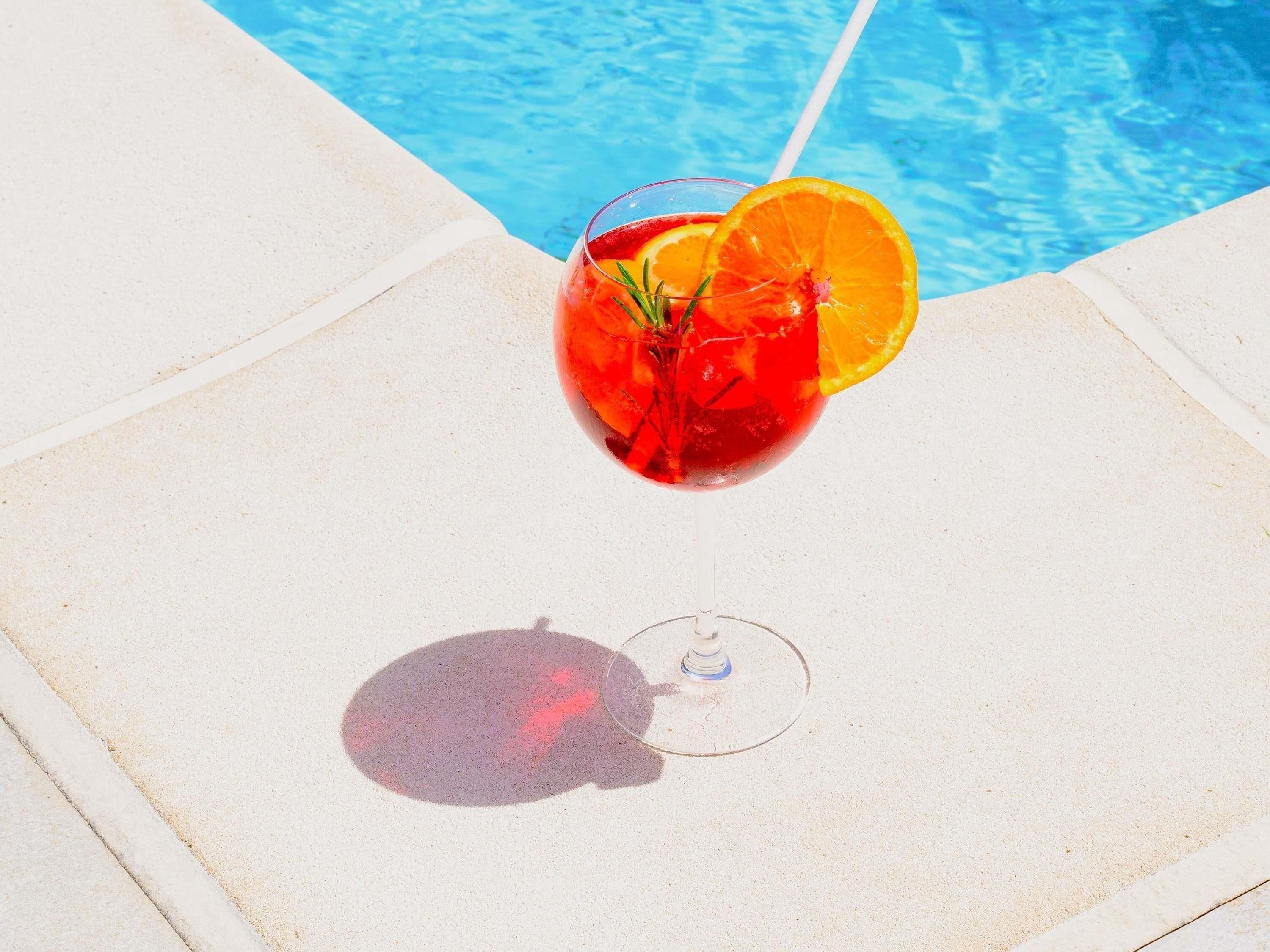 Un vaso de bebida roja con una rodaja de naranja y una ramita de romero junto a una piscina.