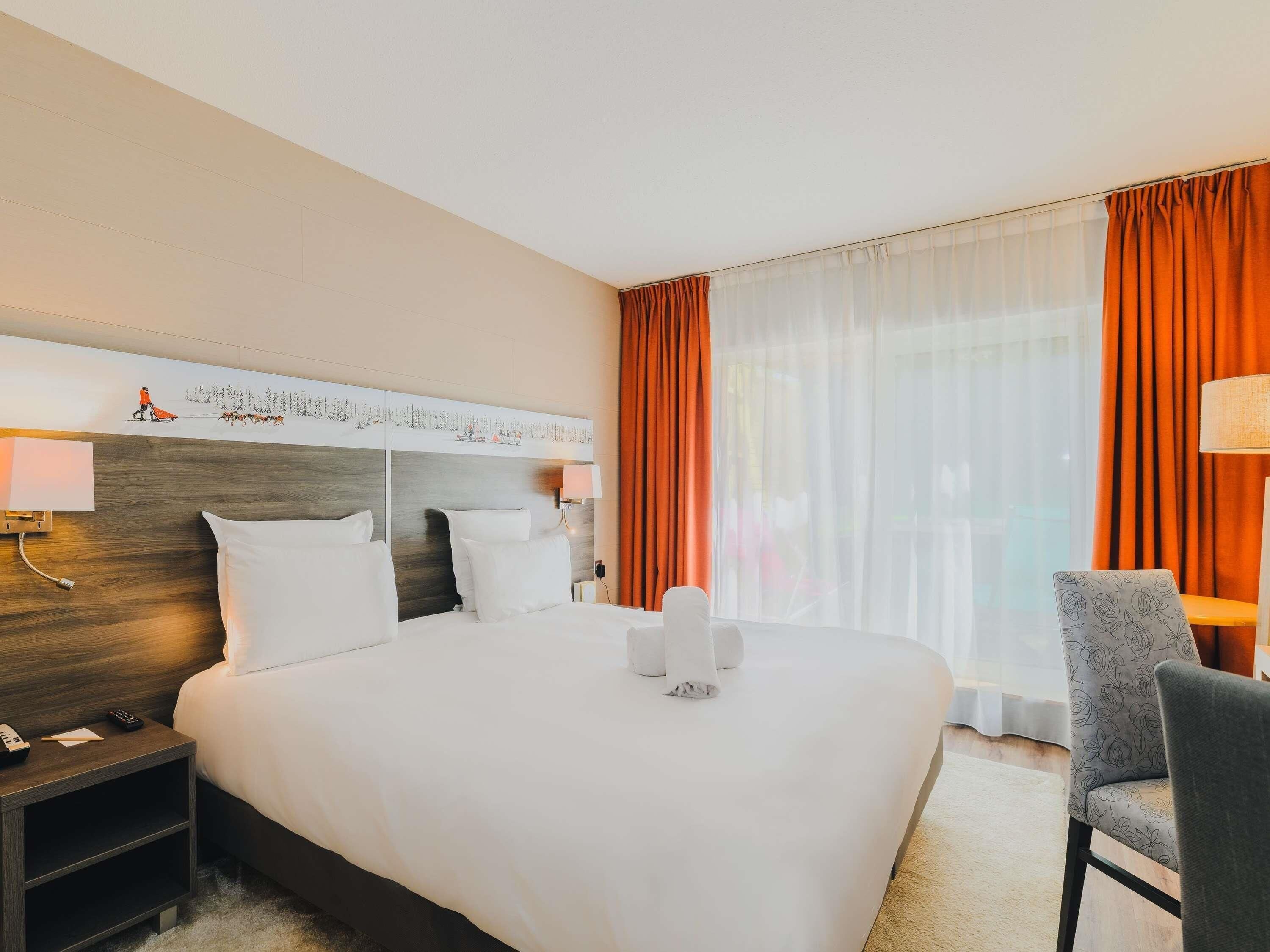 Une chambre d’hôtel avec un grand lit, une table de chevet, une chaise, une fenêtre avec des rideaux et une tête de lit ornée d’un tableau.