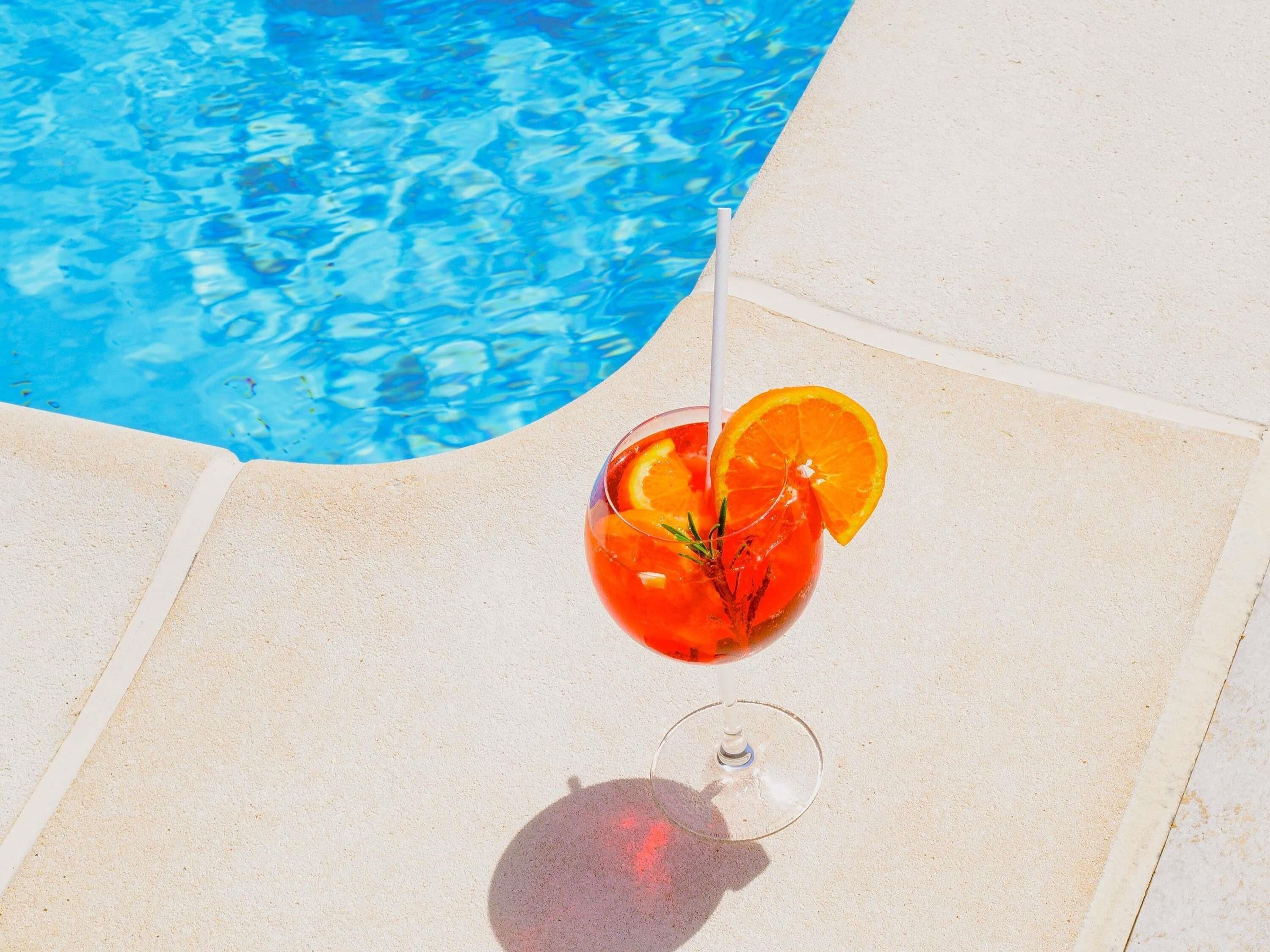 A glass of red drink with orange slices and a straw by a poolside.