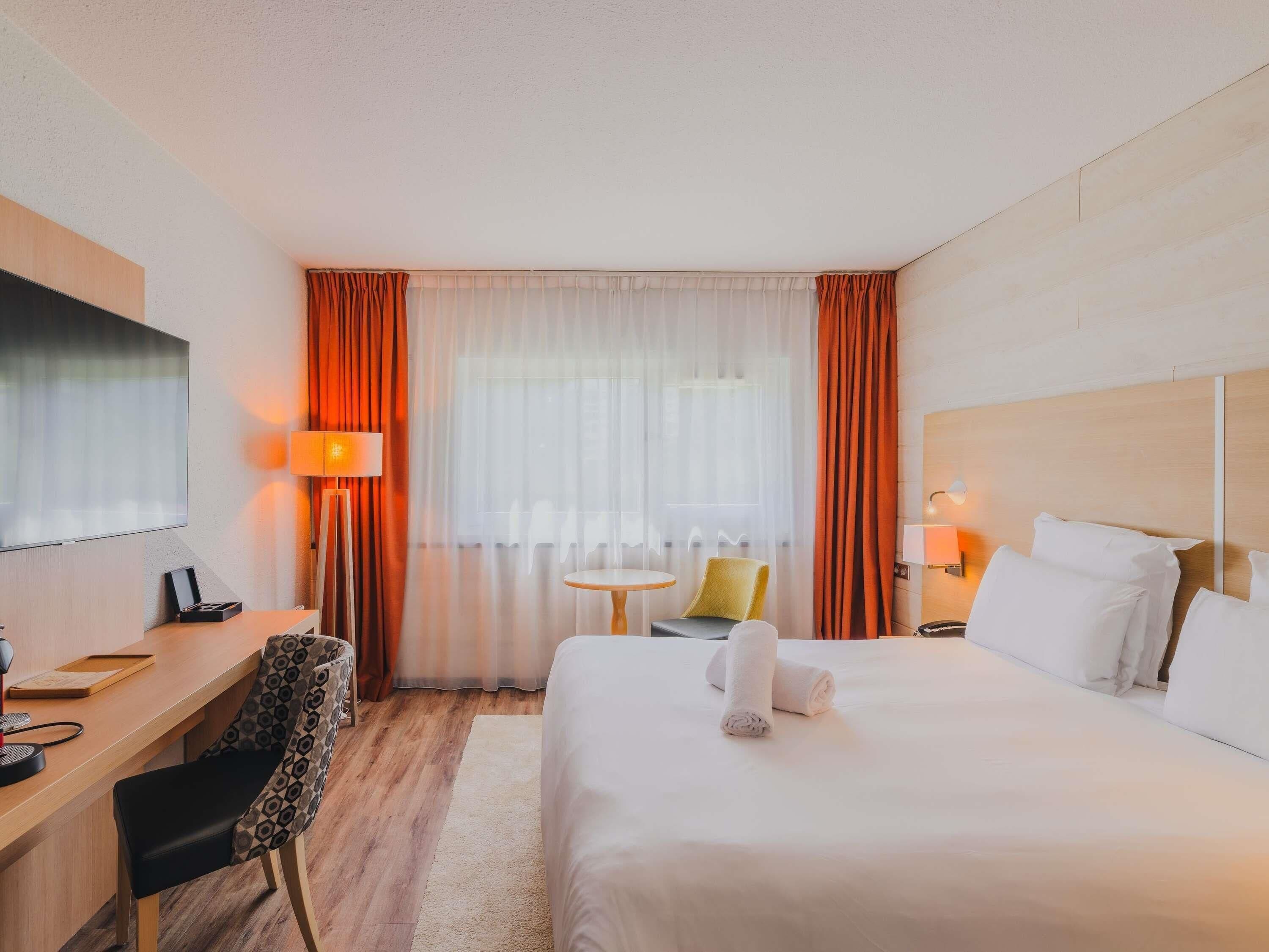 A hotel room with a bed, a desk, a chair, a TV, and a window with curtains.