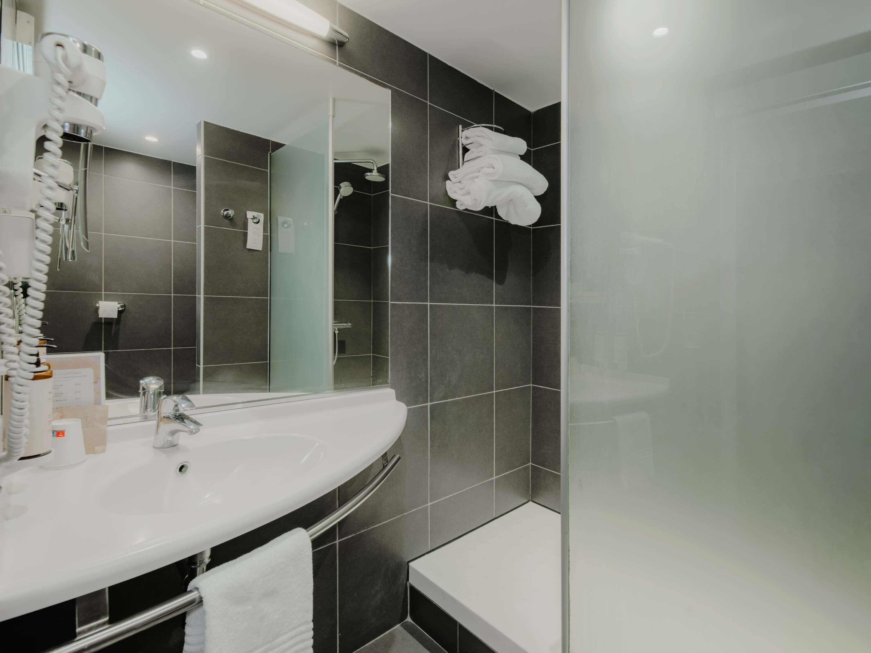 A modern bathroom with a large mirror, white sink, and dark tiled walls.