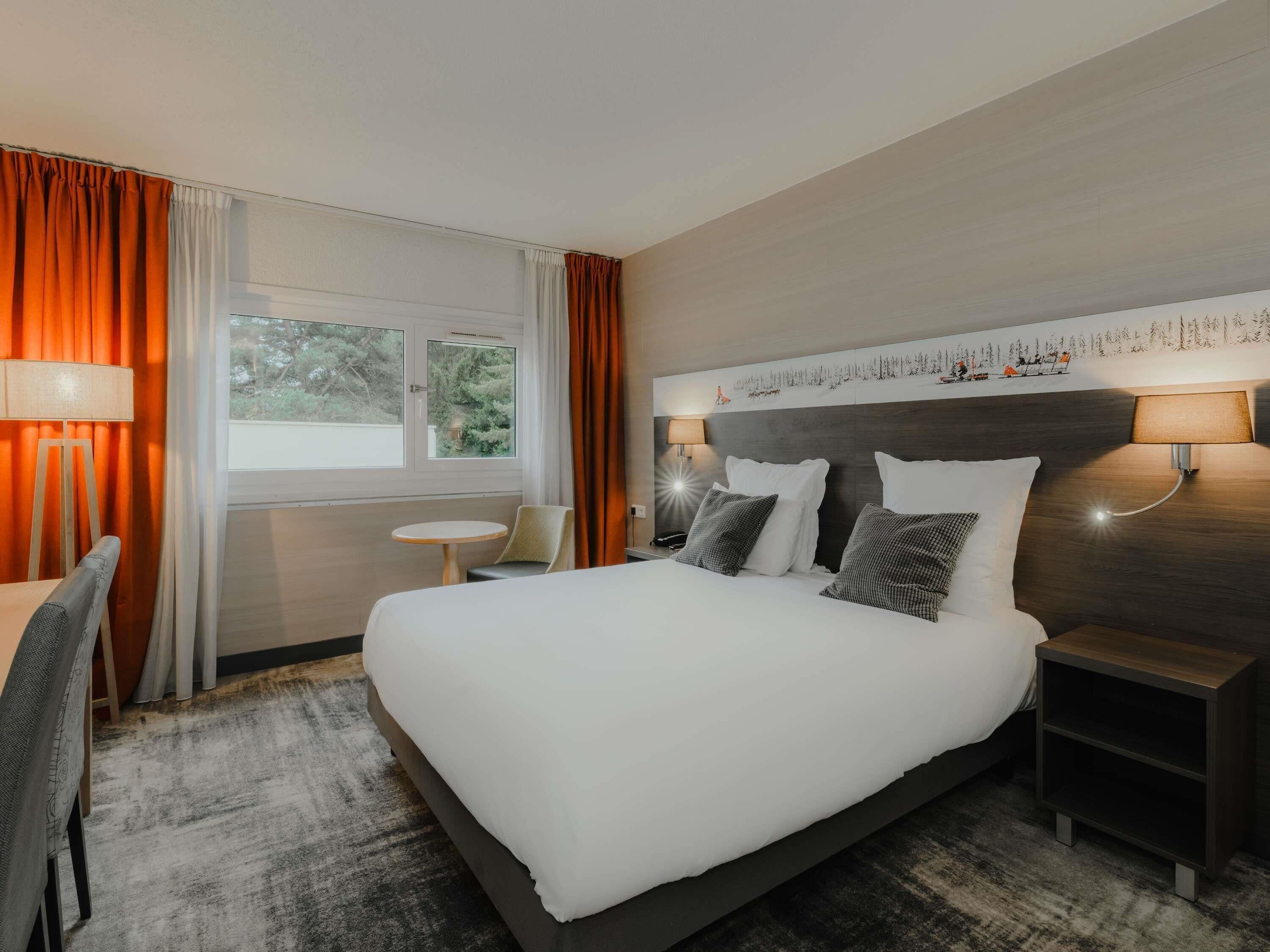 Una habitación de hotel con una cama grande, dos lámparas de noche, una mesita pequeña y una silla.