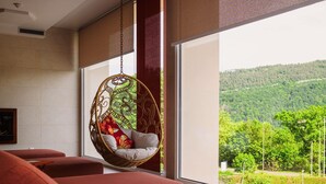 A modern interior with a hanging chair by a large window, red sofas, and a view of a green landscape. - Mercure Annecy Sud (Annecy)