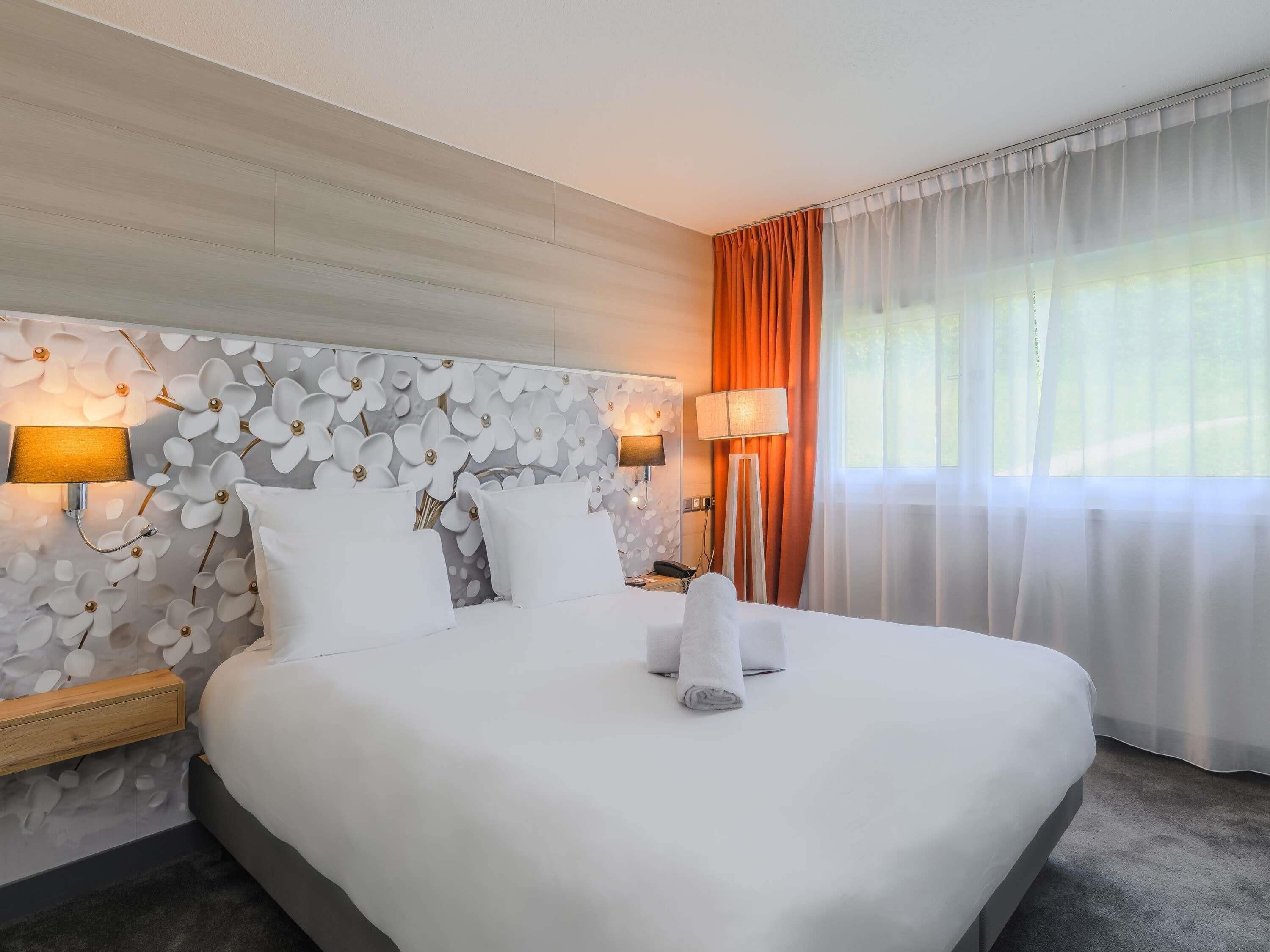 A hotel room with a large bed, two bedside lamps, a nightstand, a lamp on the floor, and a window with curtains.