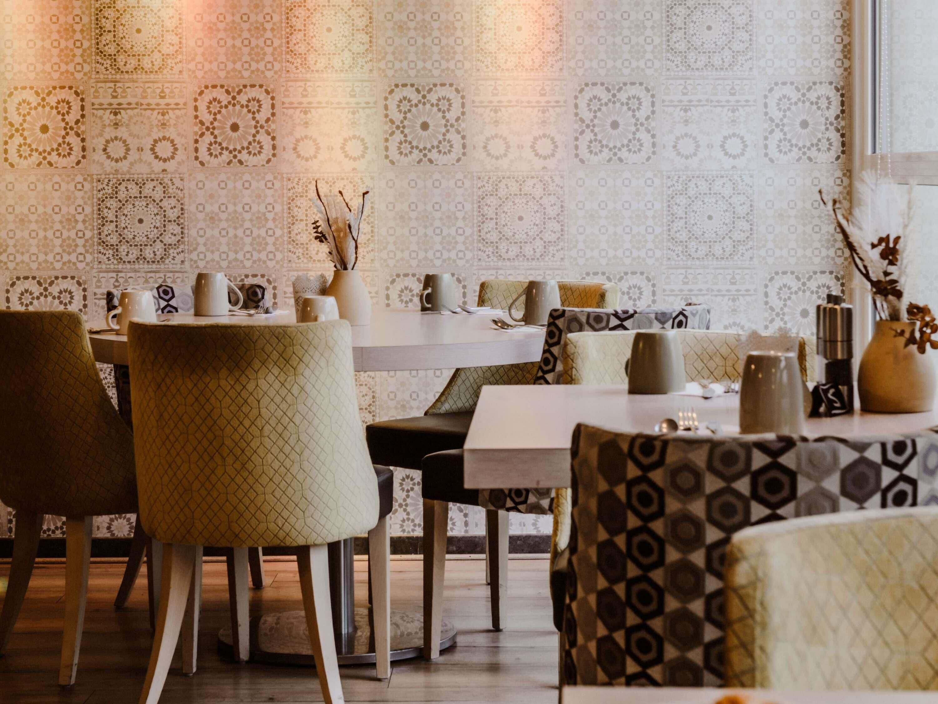 Interior de restaurante con papel tapiz estampado, mesas redondas y sillas acolchadas.