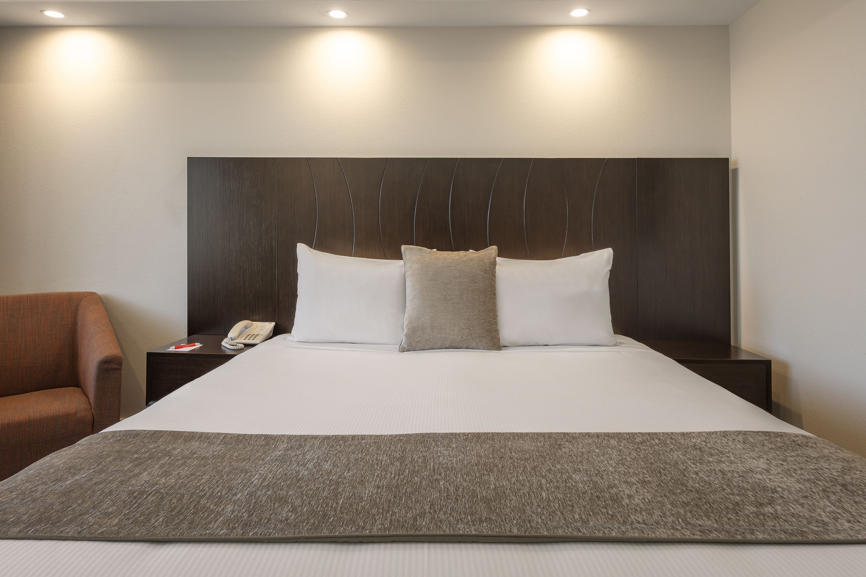 A hotel room with a large bed, a wooden headboard, a telephone on the nightstand, and a brown armchair.