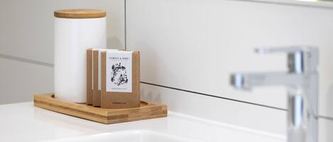 Un baño con un lavamanos blanco, una bandeja de madera que contiene un gorro de ducha y una caja con la etiqueta 'FOREST & BIRD'.