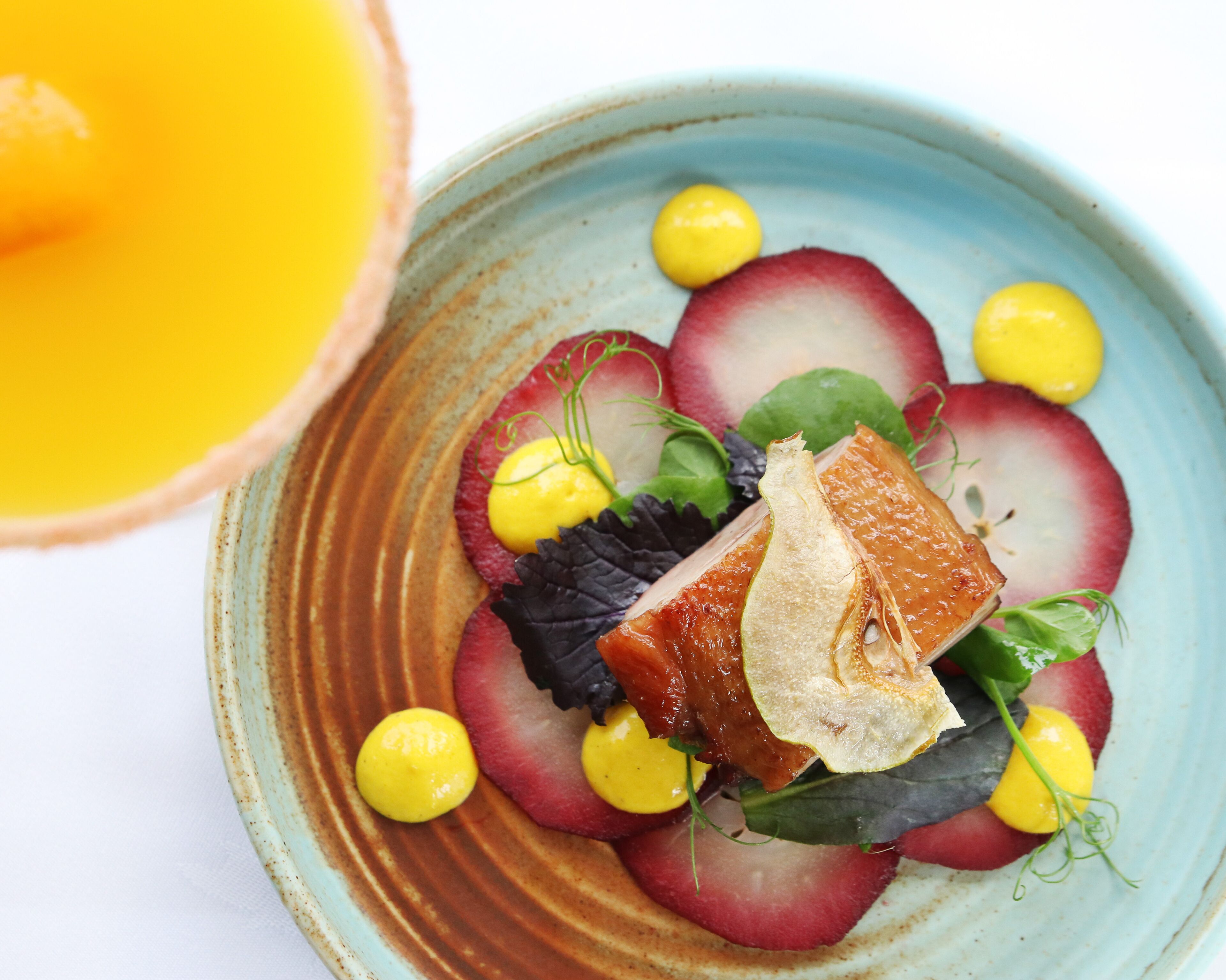 A plate with a dish of radishes, greens, and a piece of meat, possibly salmon, on a ceramic plate.