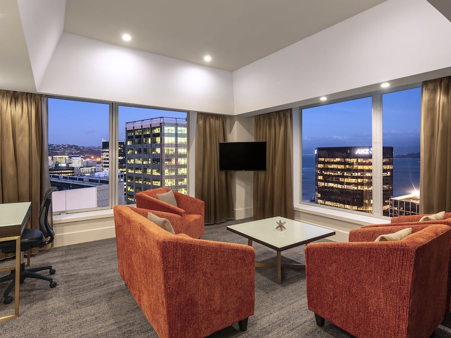 Habitación de hotel con ventanas amplias, televisor de pantalla plana, sillones tapizados en naranja, una mesita y suelo alfombrado.