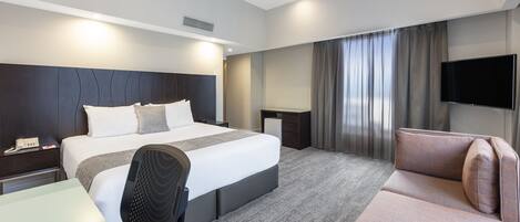 A modern hotel room with a large bed, a desk, a chair, a sofa, a flat-screen TV, and a large window with curtains.