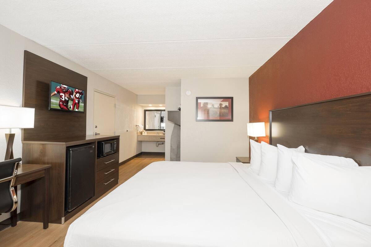 Quarto de hotel com uma cama, uma escrivaninha, micro-ondas, frigorífico, televisão e um banheiro visível ao fundo.