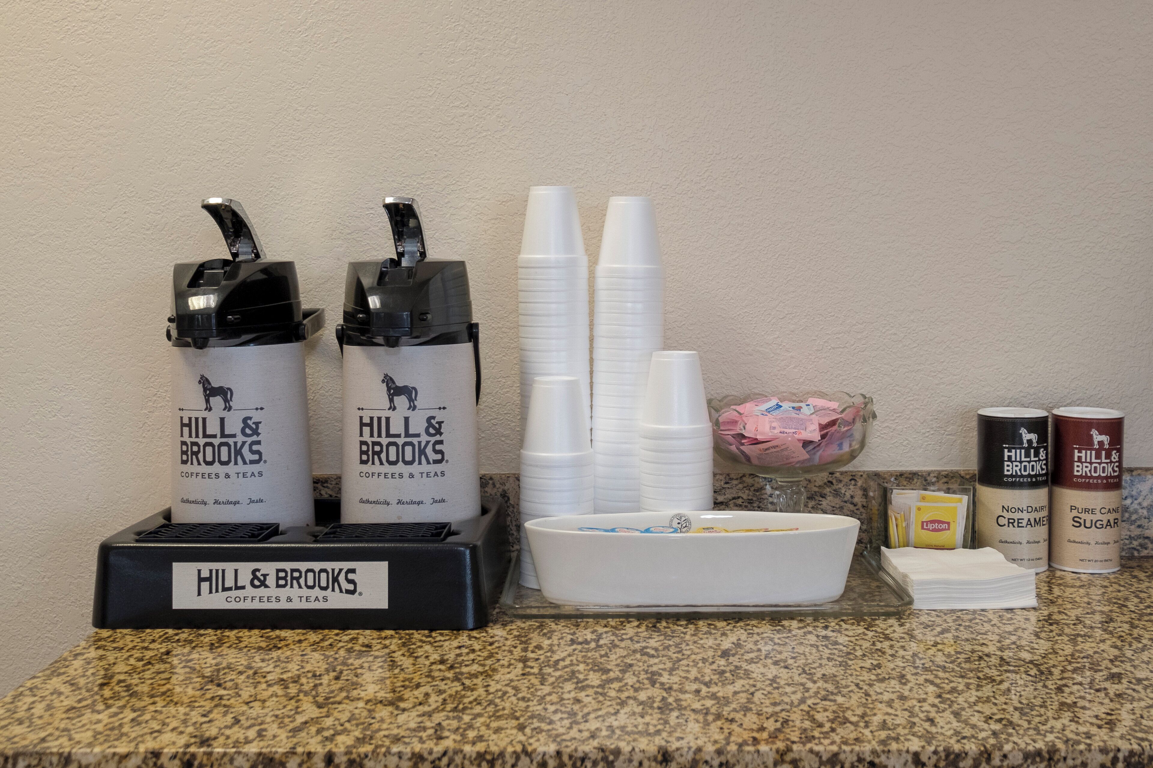 A coffee station with two dispensers, a stack of white cups, a bowl with napkins, and containers of creamer and sugar.