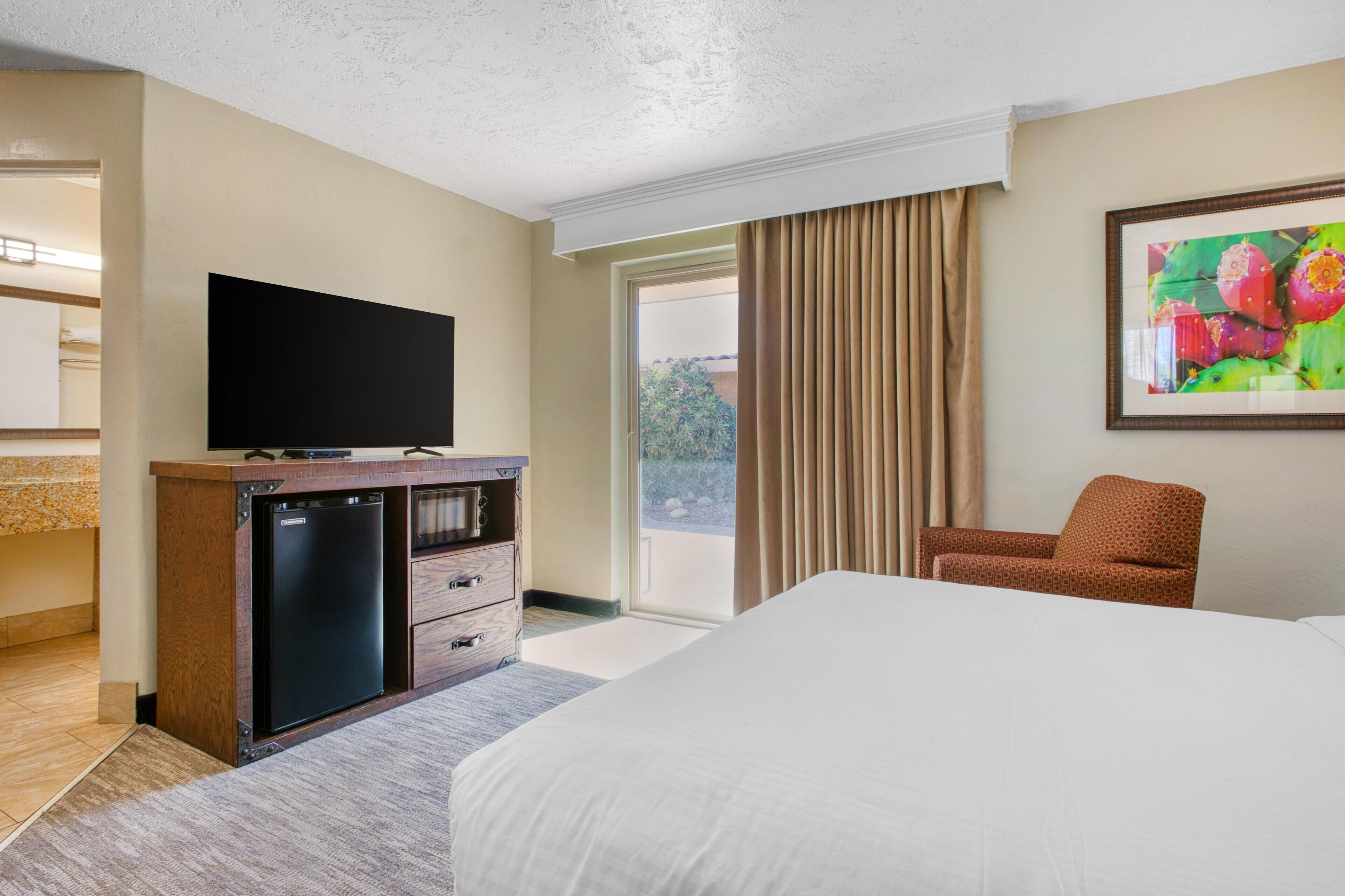 Habitación de hotel con una cama, un televisor sobre un mueble de madera, una silla y un ventanal con cortinas.