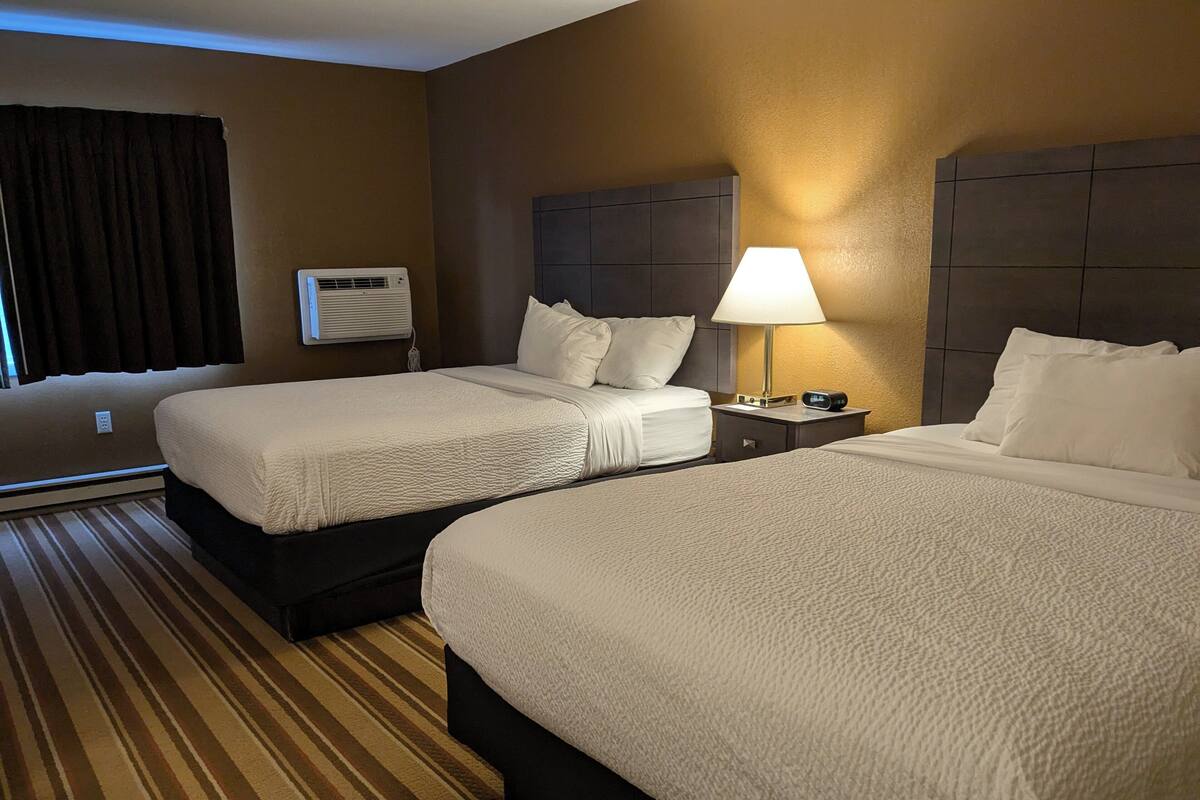 A hotel room with two beds, a bedside table with a lamp, a clock, and a window with curtains.