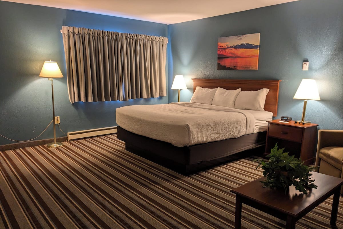 A hotel room with a large bed, two bedside lamps, a nightstand, a wooden table with a plant, and a painting on the wall.
