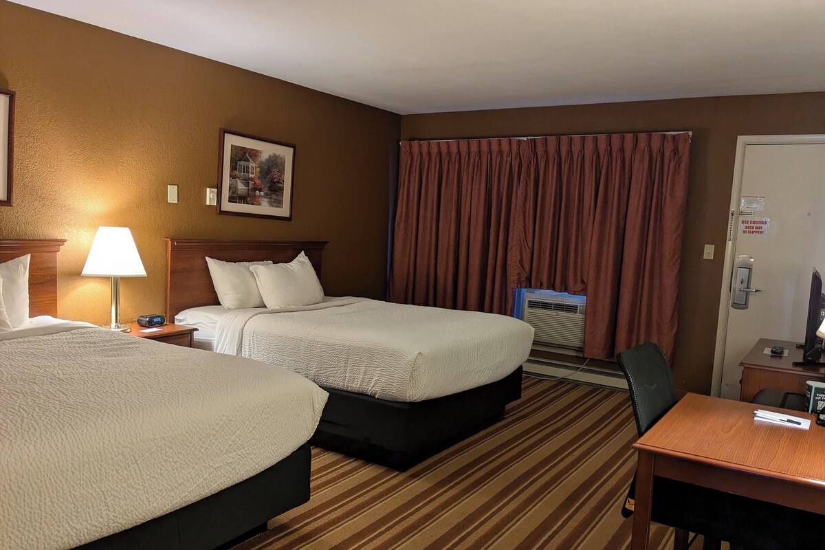 A hotel room with two beds, a desk, a chair, a lamp, a picture on the wall, and a door with a sign.