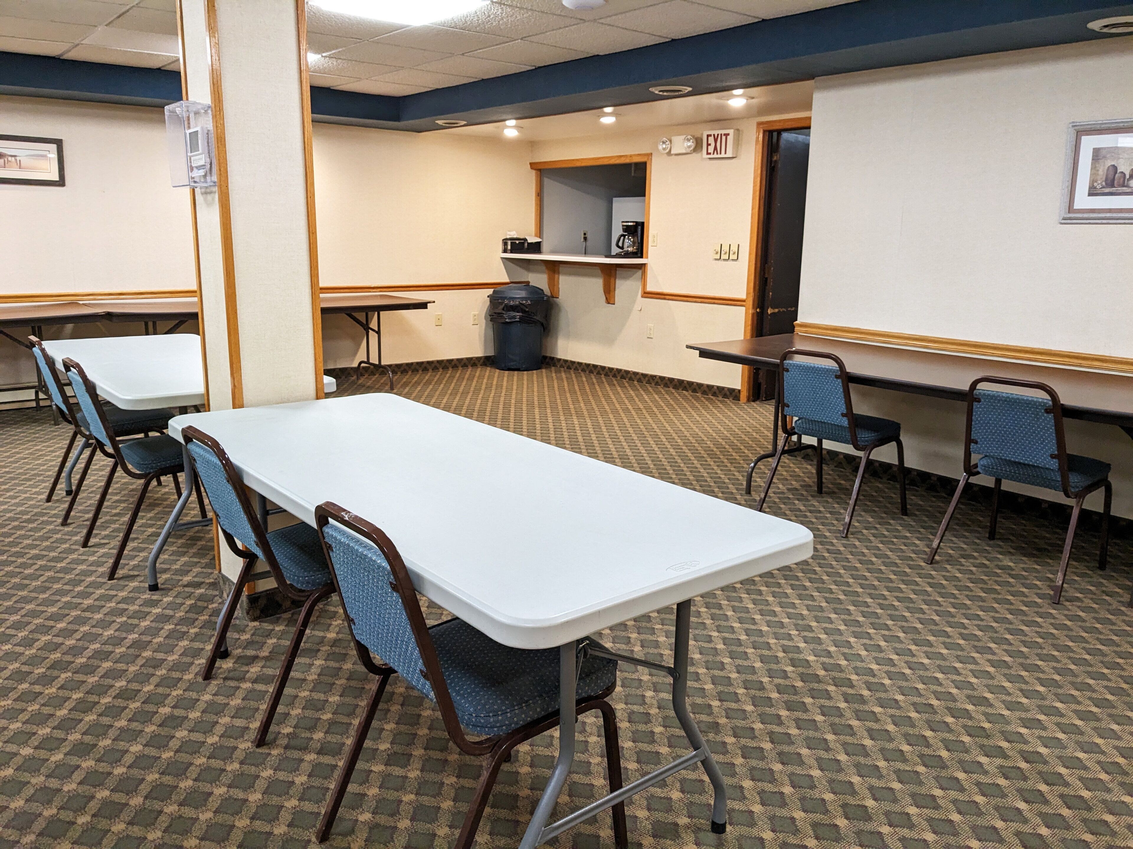 A conference room with a long table, several chairs, a trash can, and an exit sign.