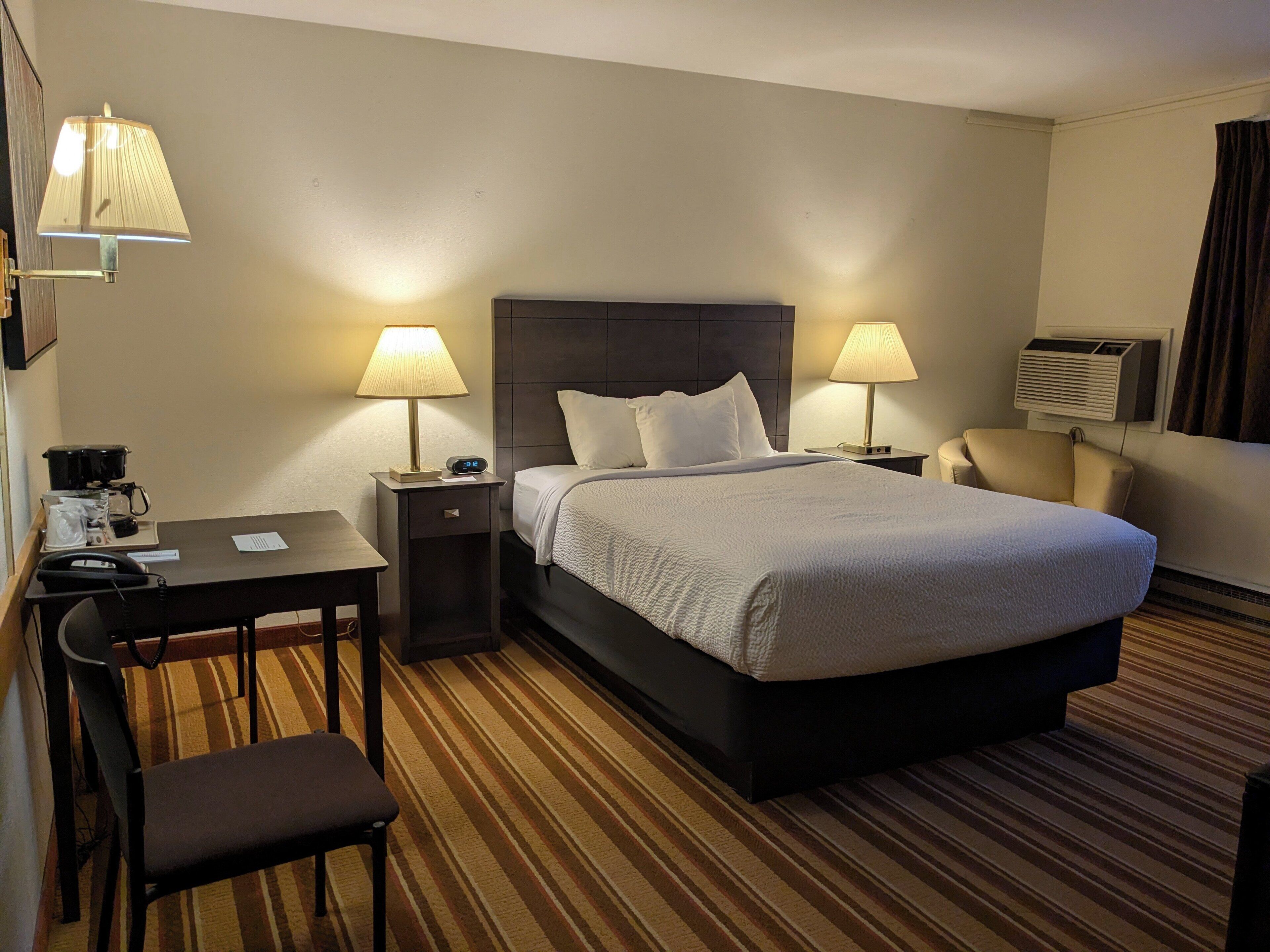 A hotel room with a bed, two bedside tables with lamps, a desk with a chair, a telephone, and an air conditioning unit.