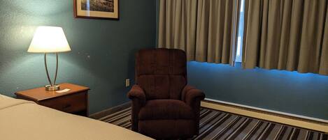 Habitación de hotel con una cama, una silla, una lámpara, un cuadro enmarcado en la pared y cortinas cubriendo una ventana.