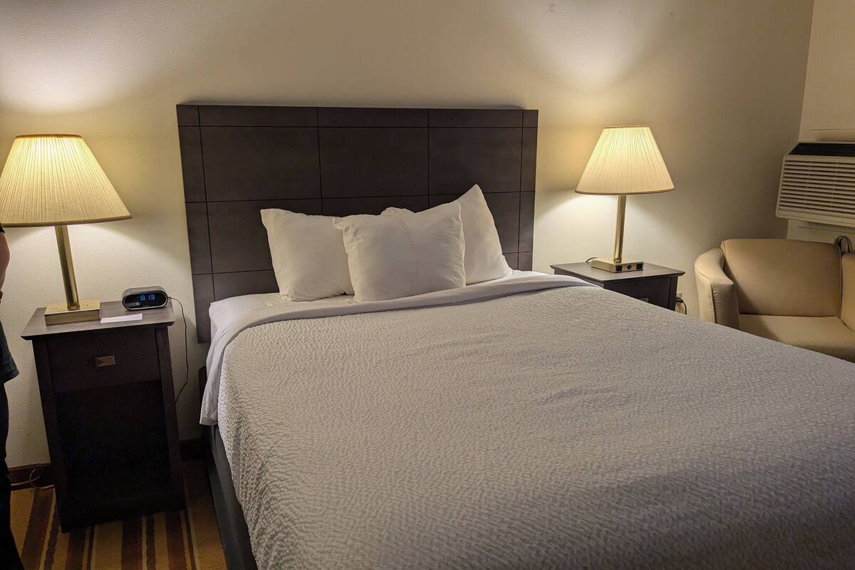 A hotel room with a large bed, two bedside lamps, a chair, and a nightstand with a clock.