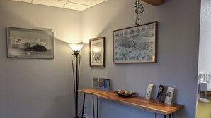 A hallway with a wooden console table displaying brochures and a fruit bowl, flanked by framed pictures and a floor lamp. - Magnuson Hotel Copper Crown (Hancock)