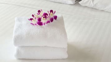 Une serviette blanche, soigneusement pliée, sur laquelle repose une orchidée aux pétales violets et blancs, le tout posé sur un lit recouvert de draps blancs.