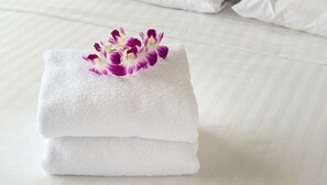 Ein ordentlich gefaltetes weißes Handtuch mit einer violett-weißen Orchidee darauf, platziert auf einem Bett mit weißer Bettwäsche.