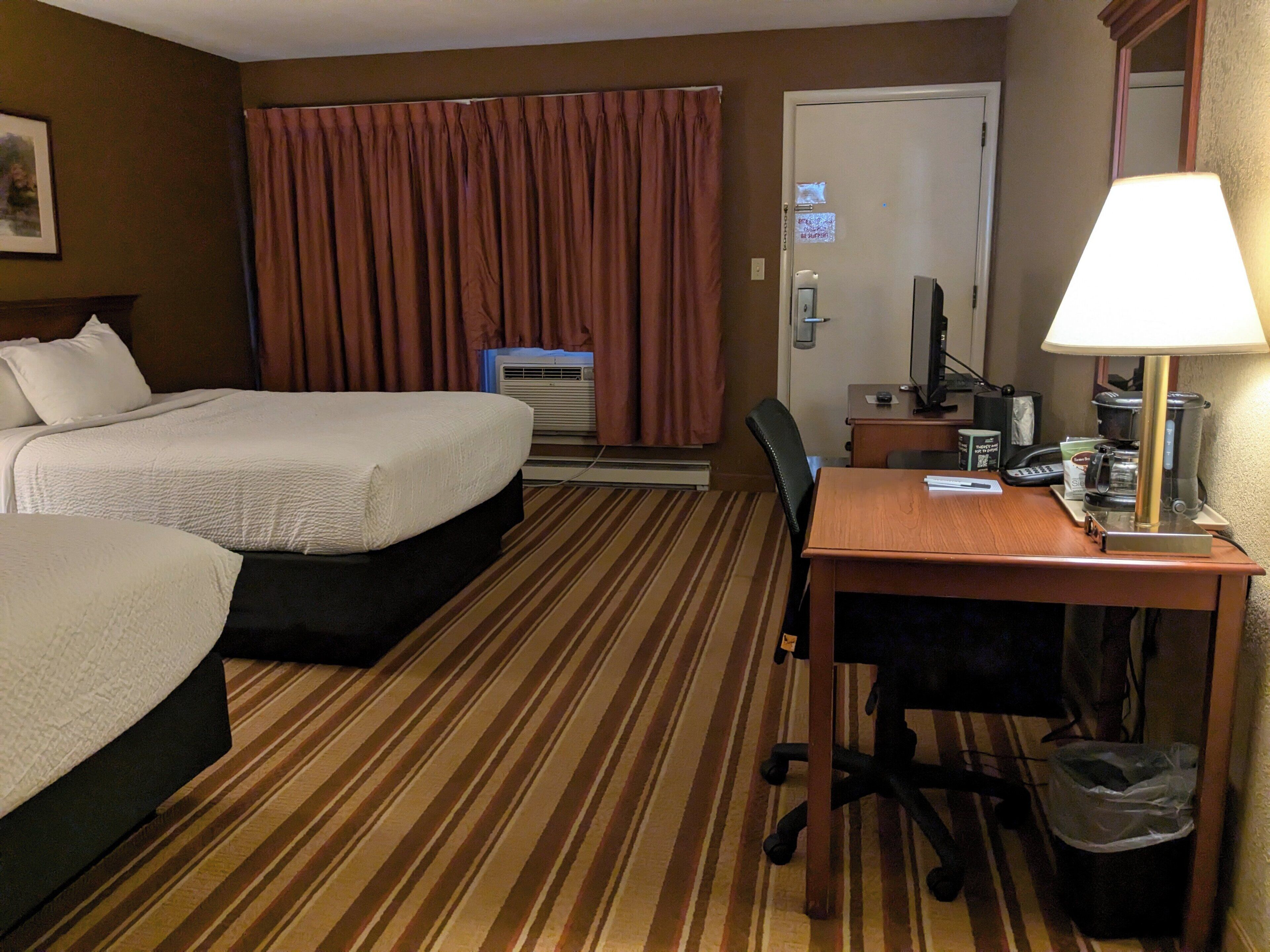 A hotel room with two beds, a desk, a chair, a lamp, a computer, and a trash bin.