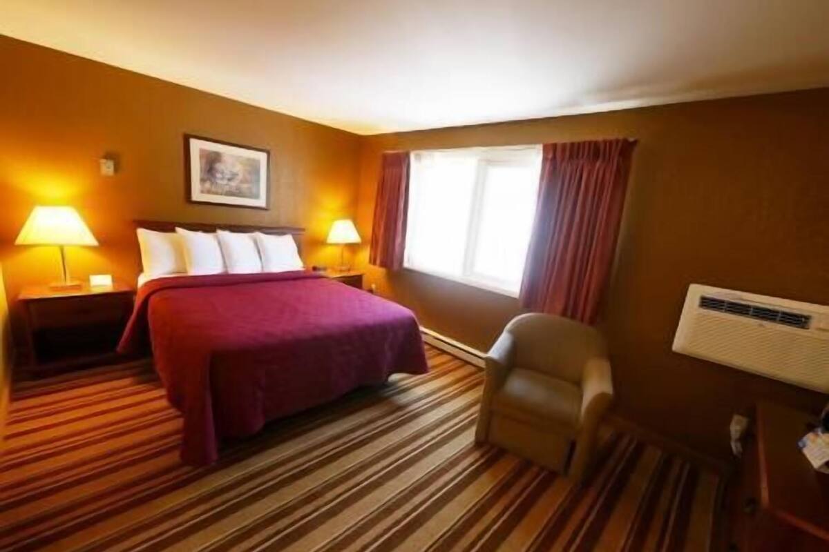 A hotel room with a bed, two bedside lamps, a chair, a window with curtains, and a wall-mounted air conditioning unit.