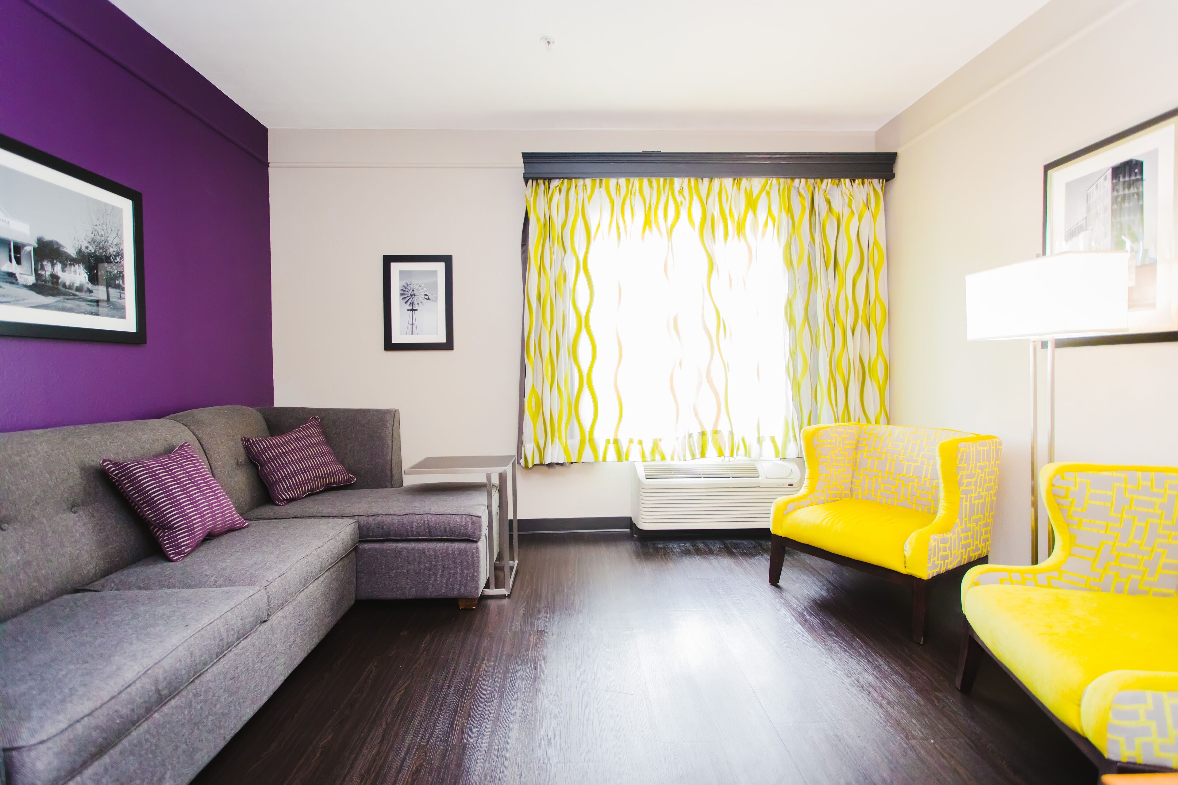A living room with a grey sofa, two yellow armchairs, a purple wall, and a window with yellow curtains.
