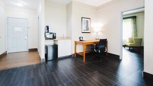 Quarto de hotel com uma escrivaninha de madeira, uma cadeira, micro-ondas, lixeira e um quadro na parede.