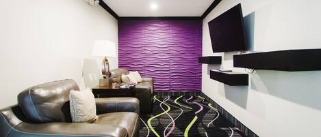 A modern hotel room with a leather sofa, a flat-screen TV mounted on the wall, a patterned carpet, and a unique purple wall design.