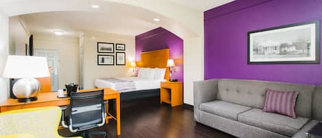 A hotel room with a bed, a desk with a lamp, a chair, a sofa, and a framed picture on the wall.