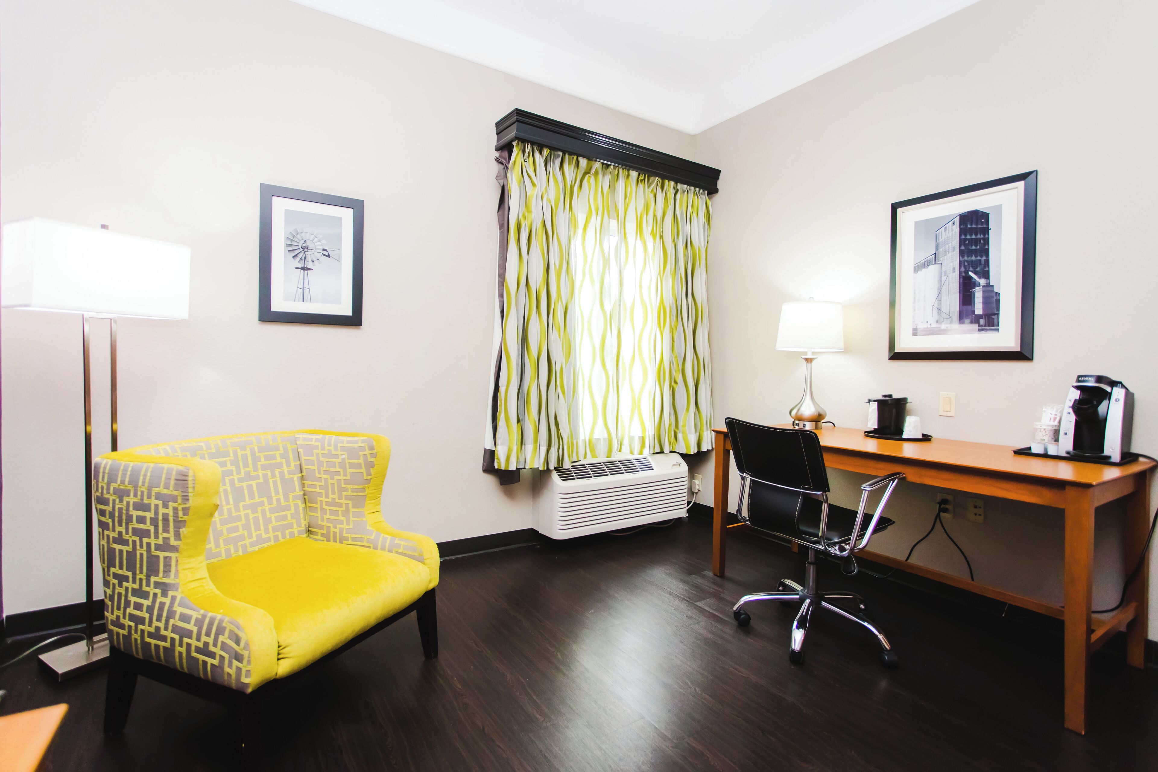 Habitación de hotel con un sillón amarillo, un escritorio de madera, una silla de oficina negra, una lámpara de pie y dos cuadros en las paredes.