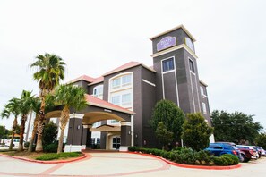 A hotel with a prominent tower, a covered entrance, and a parking lot with several cars. - Palacio Royale Inn Signature Katy (Katy)