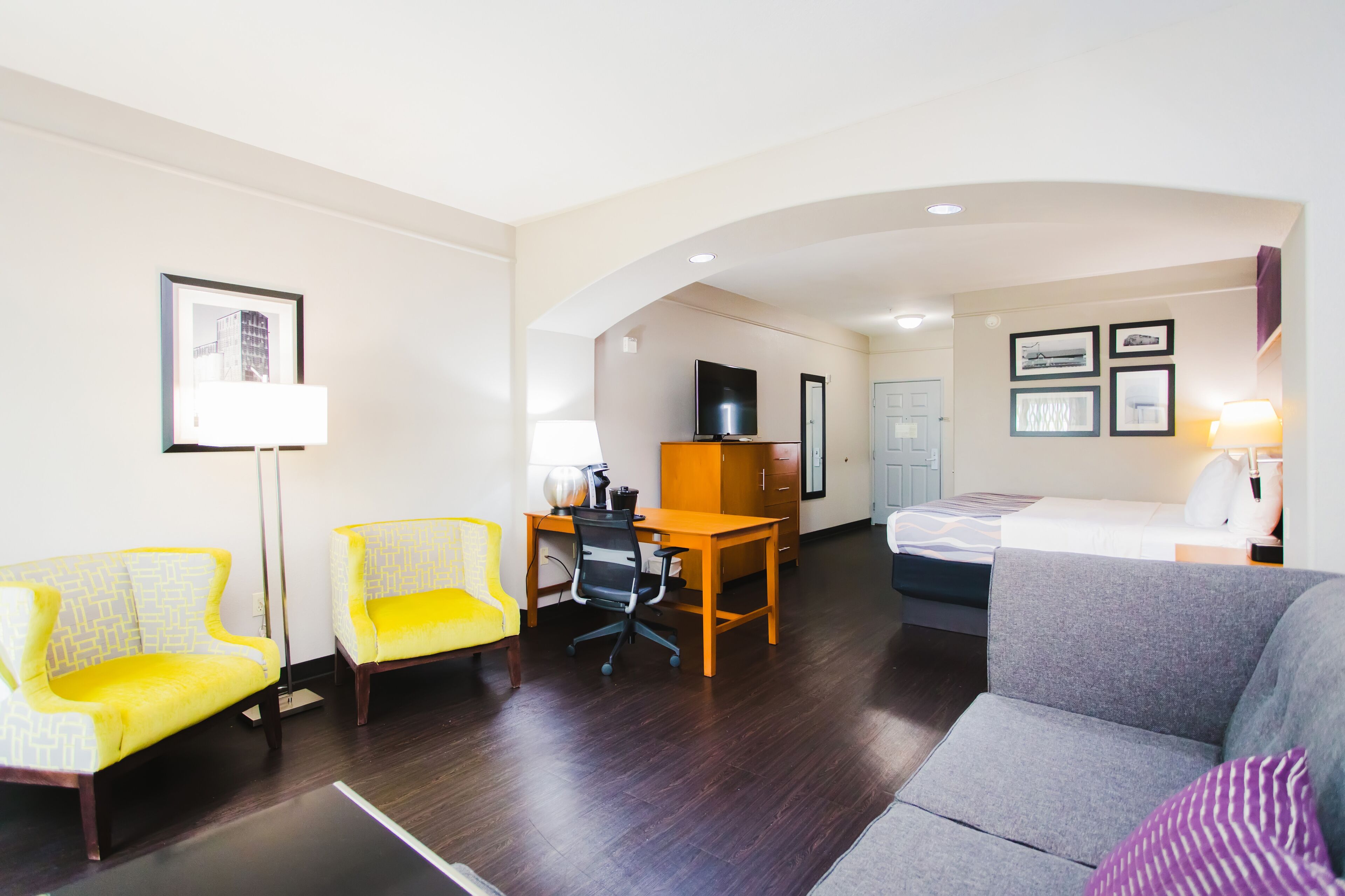 A hotel room with a sofa, two yellow armchairs, a desk with a computer, a bed, and a TV.