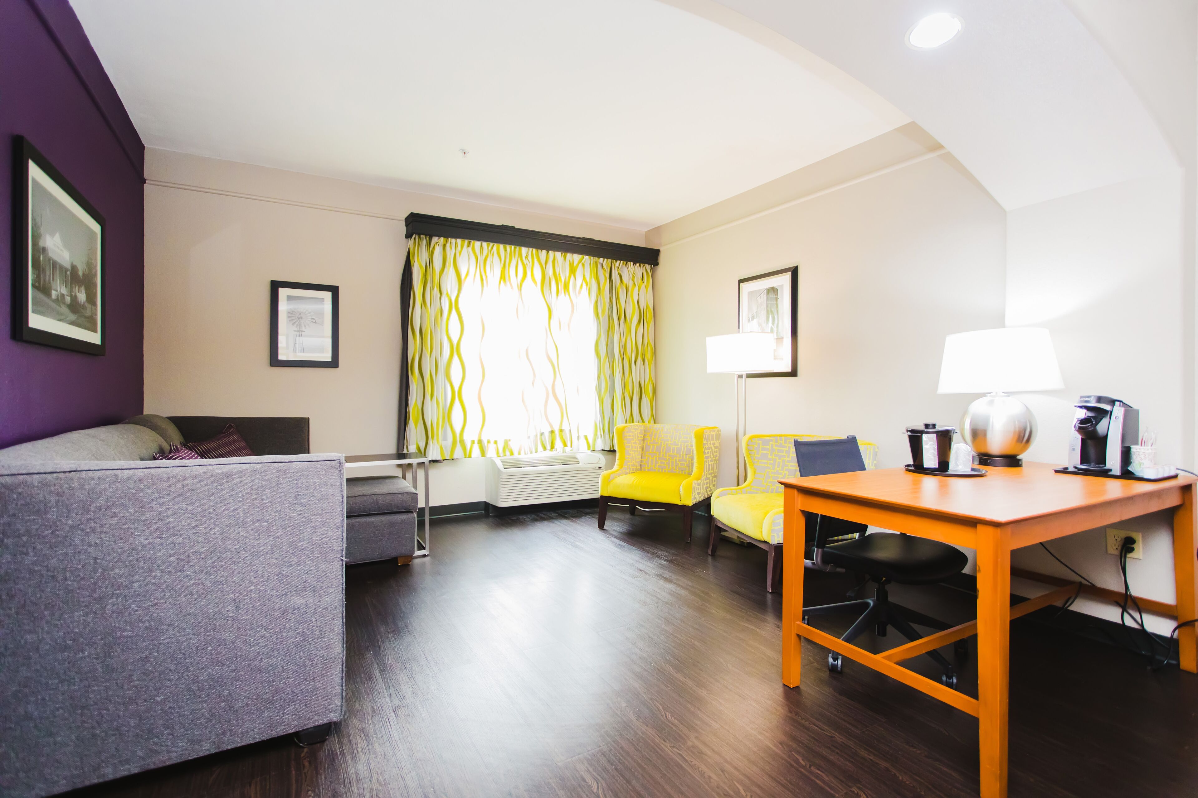Une chambre d’hôtel avec un canapé gris, un fauteuil jaune, une table en bois avec une lampe et une fenêtre avec des rideaux.