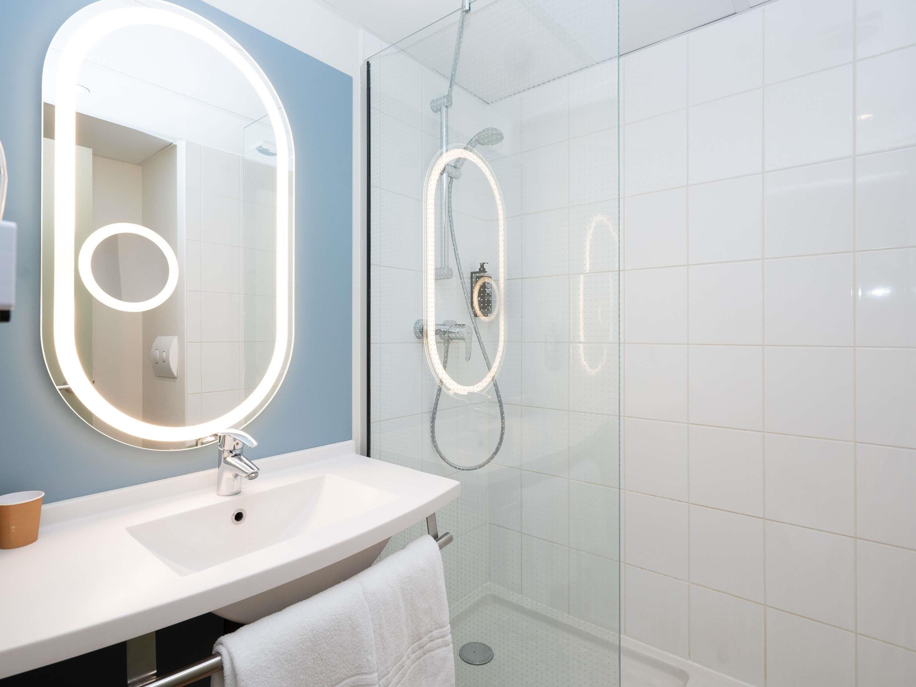 Un baño moderno con un lavamanos blanco, un espejo grande con luces integradas, una ducha con mampara de cristal y un perchero para toallas.