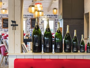 Una fila de botellas de vino sobre una barra en un restaurante, con un banco rojo y luces colgantes al fondo.