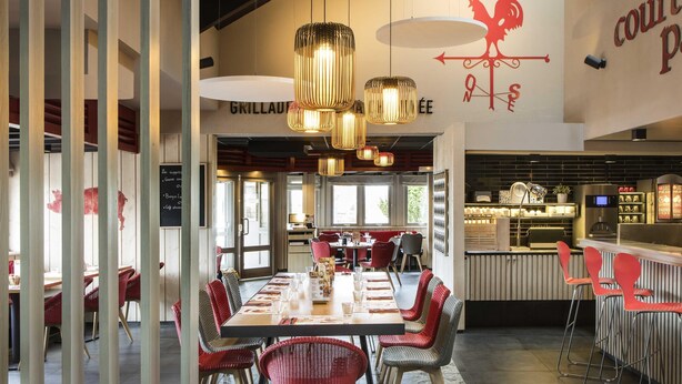 A restaurant with a long communal table, red and grey chairs, hanging pendant lights, and a wall with a rooster logo. - ibis Reims Tinqueux (Tinqueux)