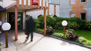 A hotel entrance with a red 'ibis' sign, a covered walkway, and a well-maintained garden with various plants and flowers. - ibis Reims Tinqueux (Tinqueux)