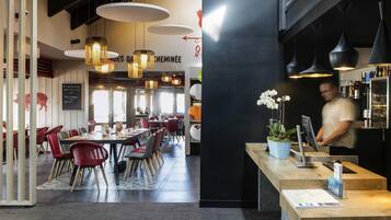 A restaurant interior with high ceilings, hanging pendant lights, and a central dining area with red chairs and a black table.