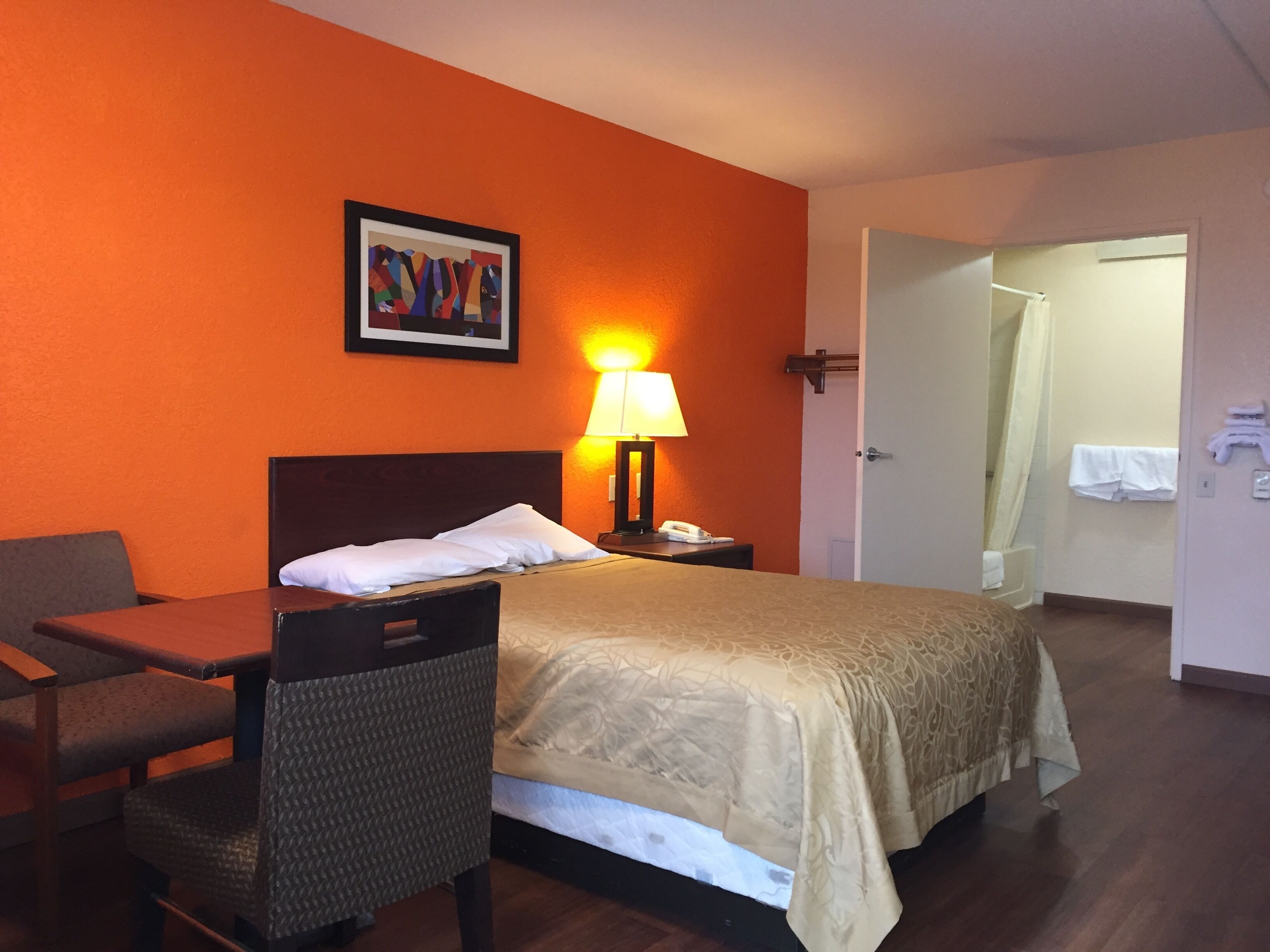 Une chambre d’hôtel avec un lit, un bureau, une chaise, une lampe et une salle de bain visible à travers une porte ouverte.