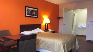 Une chambre d’hôtel avec un lit, un bureau, une chaise, une lampe et une salle de bain visible à travers une porte ouverte.