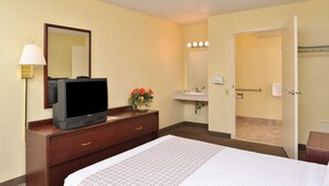 Ein Hotelzimmer mit Bett, Fernseher auf einem Schminktisch, Spiegel, Waschbecken und einer Tür zu einem weiteren Raum.