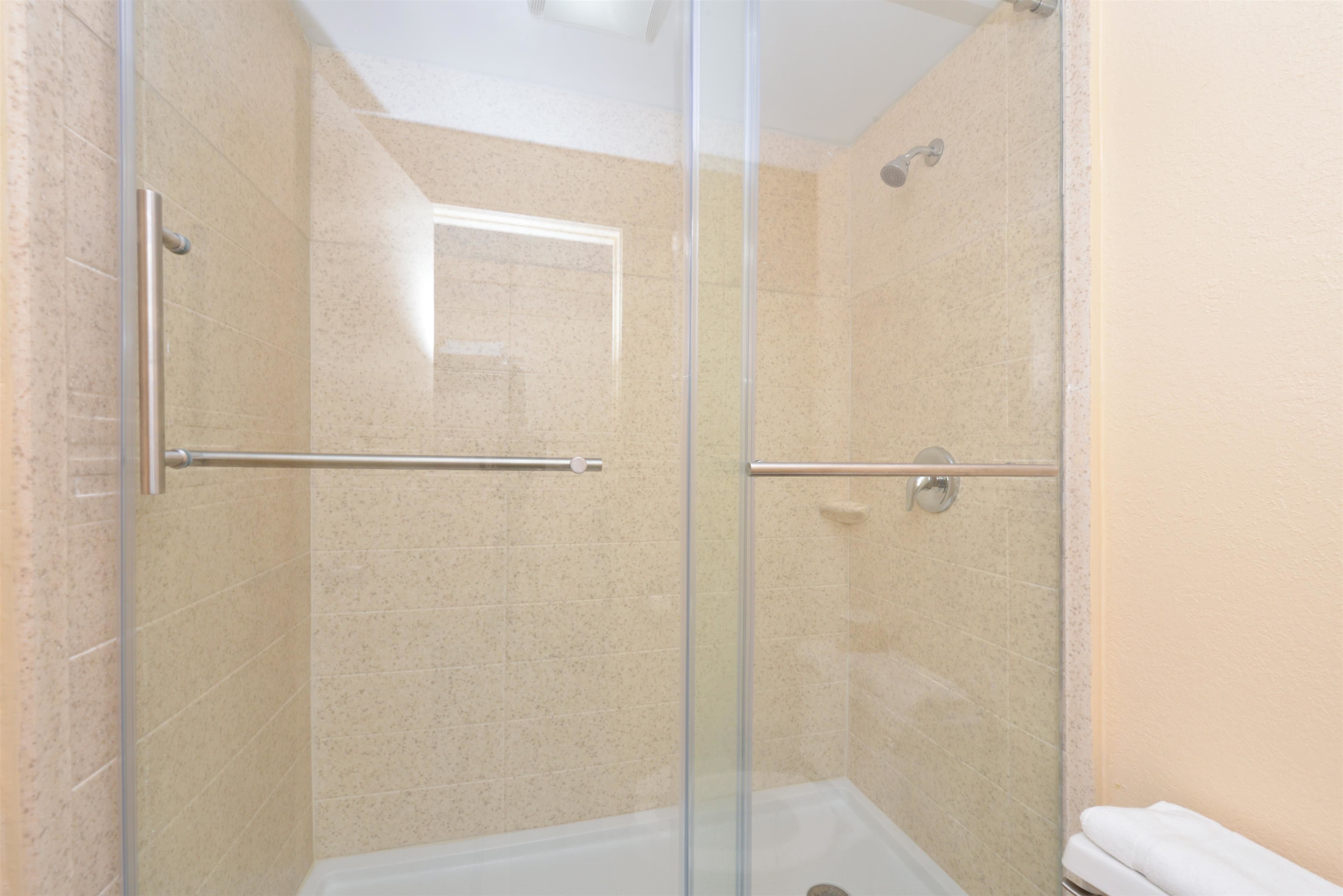 Una ducha con cerramiento de vidrio, barra de metal, paredes de azulejos color beige y una bañera blanca.