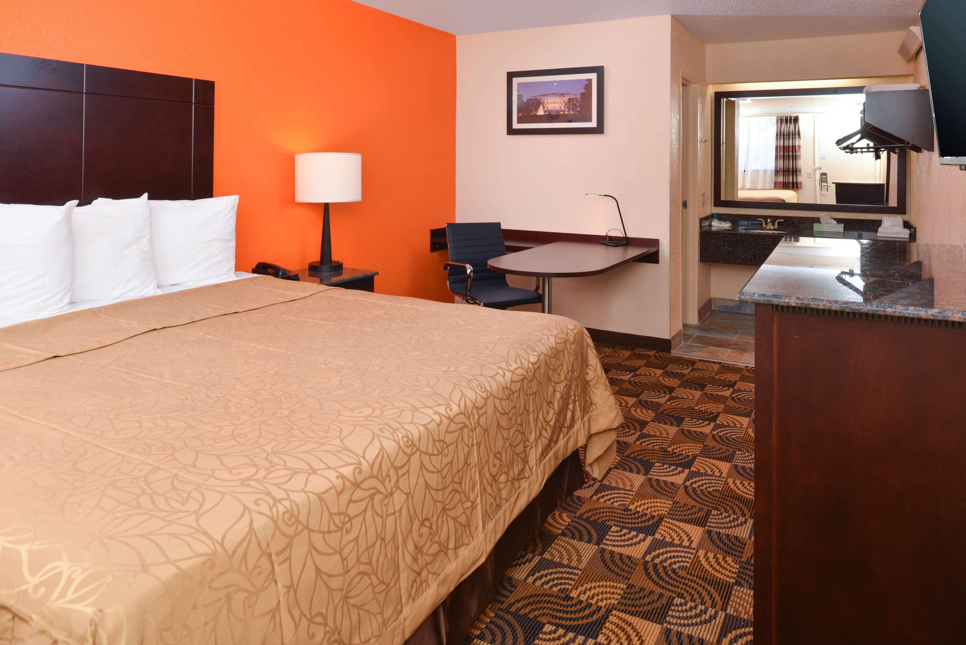 A hotel room with a bed, a desk, a chair, a desk lamp, a mirror, and a bathroom visible through an open door.