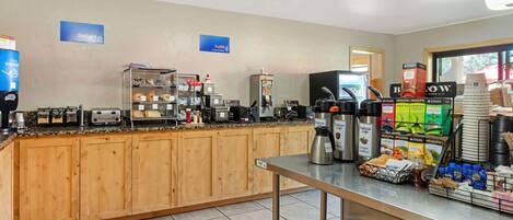 Un área de desayuno autoservicio con diversas opciones de comida y bebida, que incluye cafeteras, un refrigerador y una selección de aperitivos y bebidas.