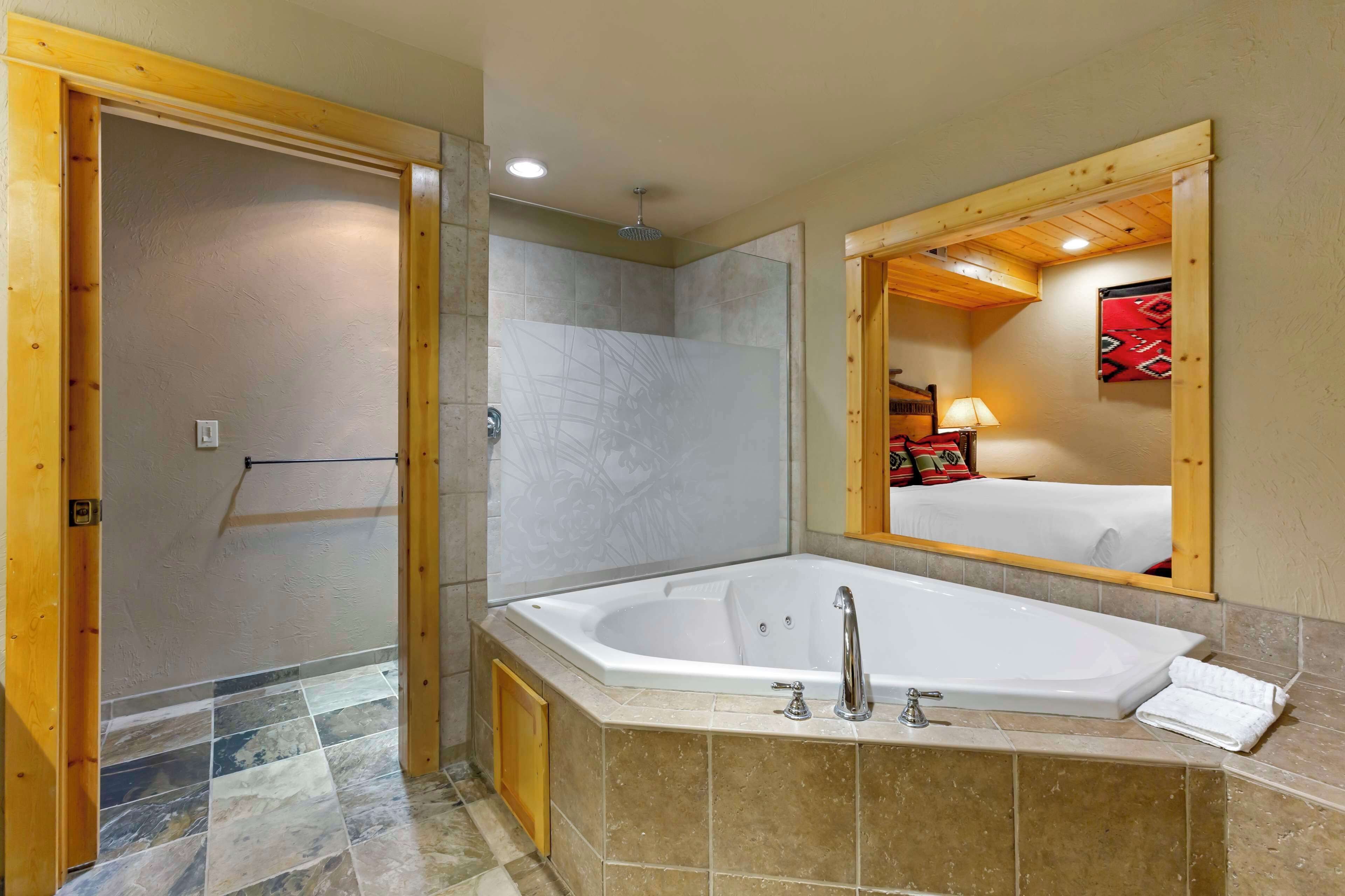 Un baño con una bañera grande, zona de ducha y un espejo que refleja una cama con una colcha roja.